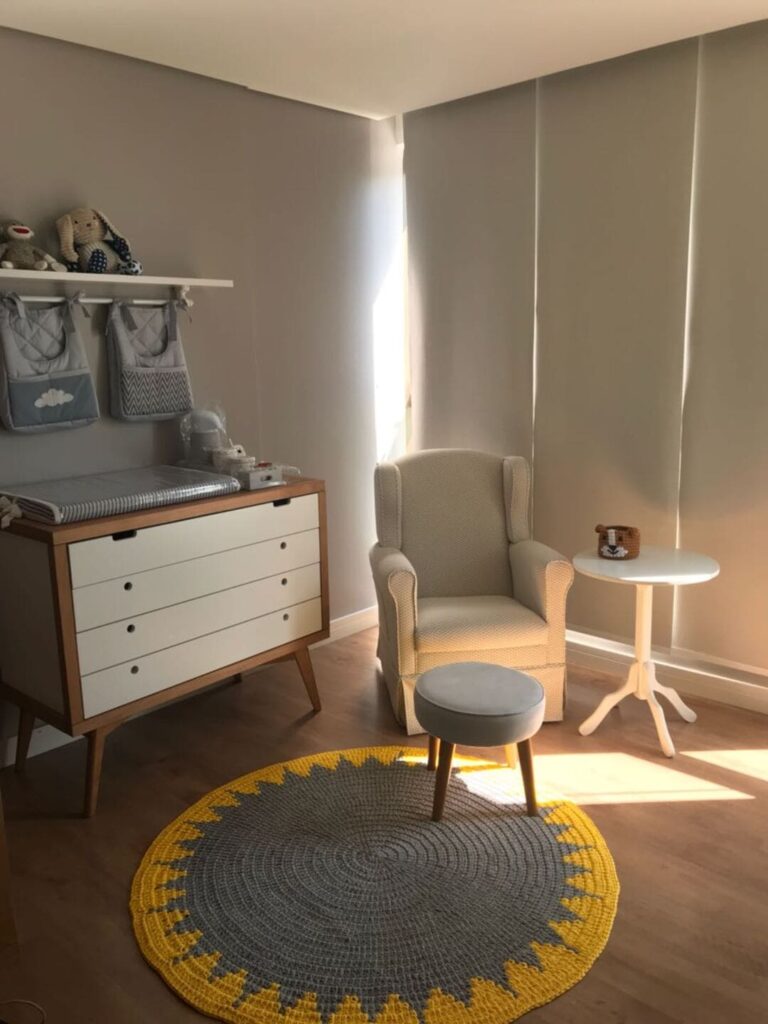 Tapete de crochê para quarto de bebê redondo, nas cores cinza e amarelo. Ao fundo, cadeira e mesa em tons claros, e gaveteiro em branco e marrom. 