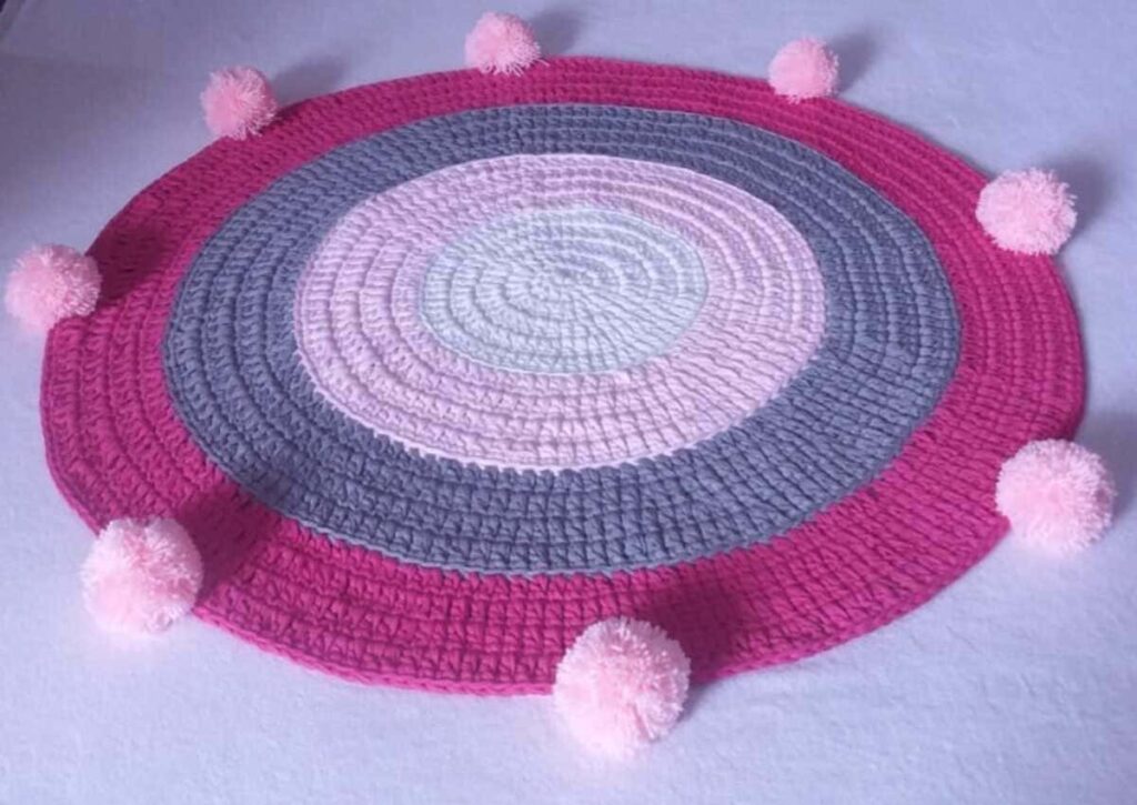 Tapete de crochê tradicional, ponto médio, redondo, nas cores rosa choque, cinza, rosa claro e branco, com pompons rosas removíveis nas extremidades