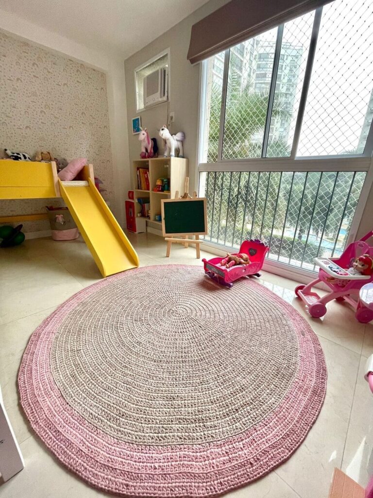 Tapete de crochê para quarto infantil redondo, em rosa e cinza, com quarto ao fundo com brinquedos e cama com escorregador amarelo