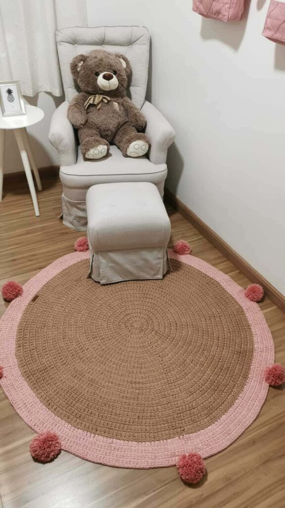 Tapete de crochê para quarto de criança redondo, em rosa e marrom, com pompons rosa removíveis. Ao fundo, poltrona cinza com urso de pelúcia