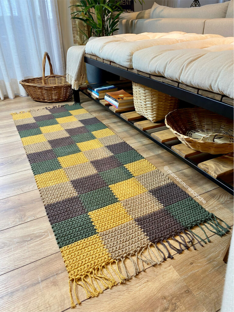 Tapete de crochê colorido, retangular, quadriculado nas cores bege, amarelo, verde e azul. Ao lado, sofá, cestos de vime e plantas decorativas