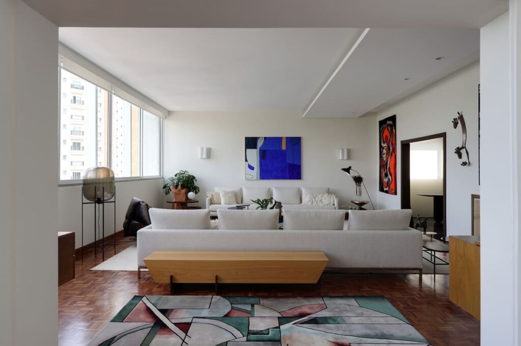 Sala de estar com sofá, quadros e janela à esquerda. À frente, tapete colorido, com desenho geométrico