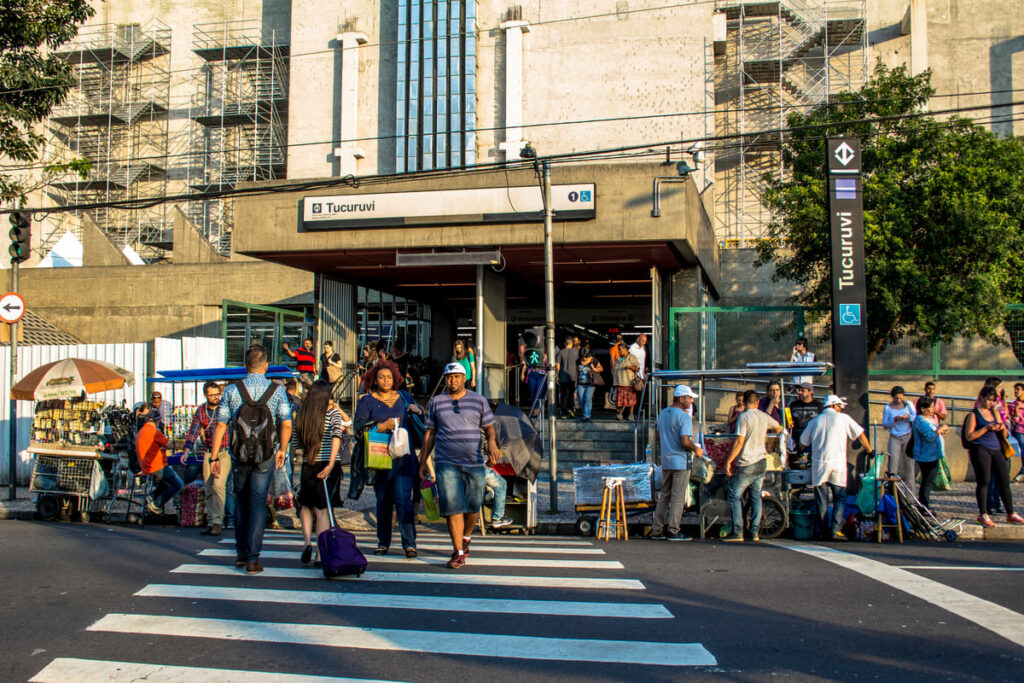 Porta da Estação Tucuruvi do Metrô de SP com pessoas na porta e pedestres atravessando a faixa em frente à entrada
