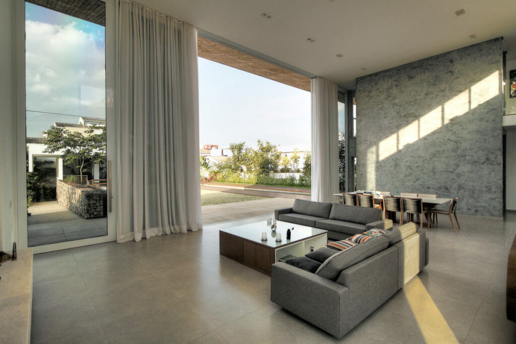 Sala ampla, com mesa e sofás, com pé direito duplo, integrada ao jardim. Porcelanato no piso e pedras naturais na parede ampliam a sensação de conforto da casa. 