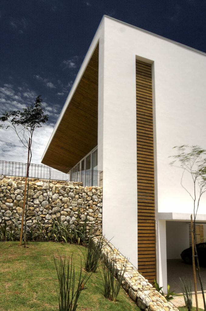 Fachada de casa branca, com acabamentos em madeira e pedras naturais e jardim em frente