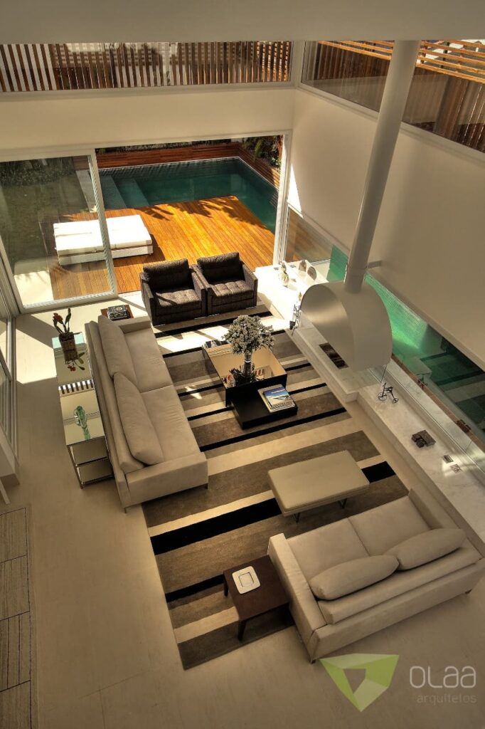 Vista de cima de sala ampla de casa de luxo, em tons claros, com sofás, poltronas e integração com a varanda e a piscina do lado de fora