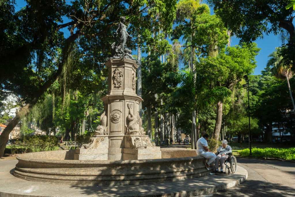 Estátua do Jardim do Palácio do Catete com duas pessoas sentadas em dia de sol com árvores ao fundo