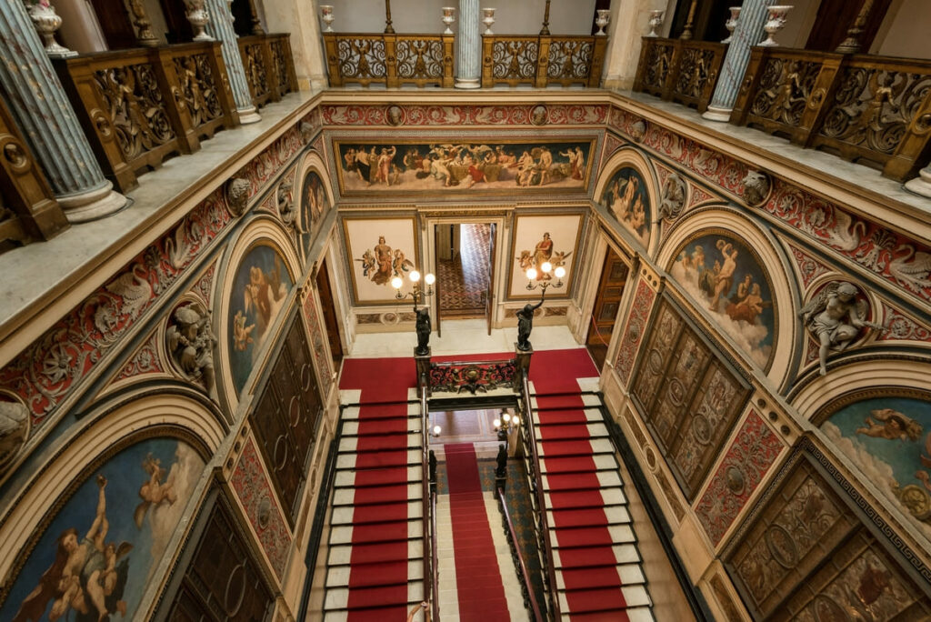 Interior do Palácio do Catete, hoje Museu da República, mostra escada com tapete vermelho, paredes totalmente decoradas com pinturas e escultas