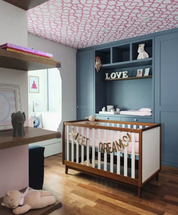 Quarto de bebê com paredes azuis escuras e papel de parede no teto em branco e rosa, com berço de madeira e bichos de pelúcia de enfreite
