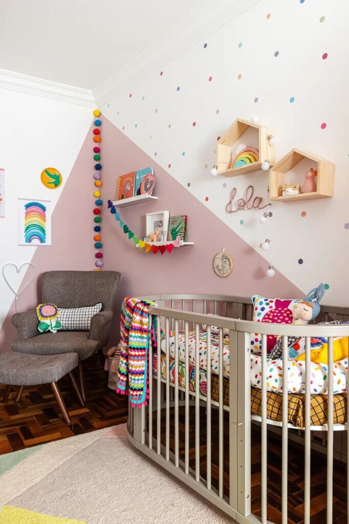 Quarto de bebê com pintura geométrica na parede e móveis em cinza, e decoração de livros e pelúcias coloridas