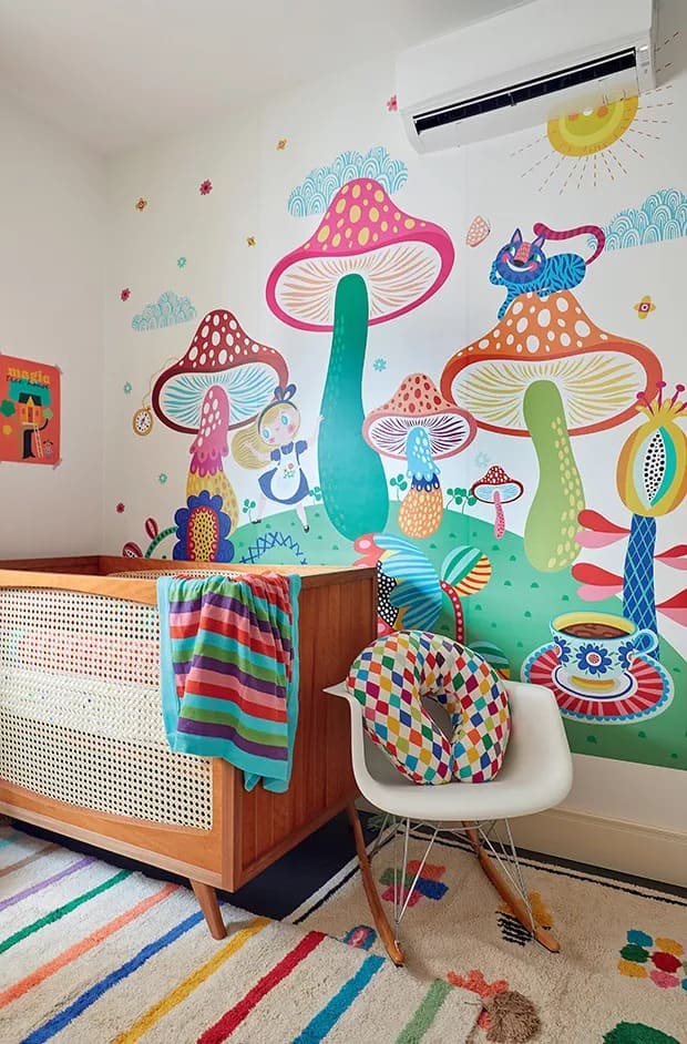 Quarto de bebê multicolorido, com papel de parede inspirado em Alice no País das Maravilhas, com imagem da Alice, cogumelos, chá e relógios. Em frente, berço em madeira e poltrona branca com almodafa com losangos coloridos 