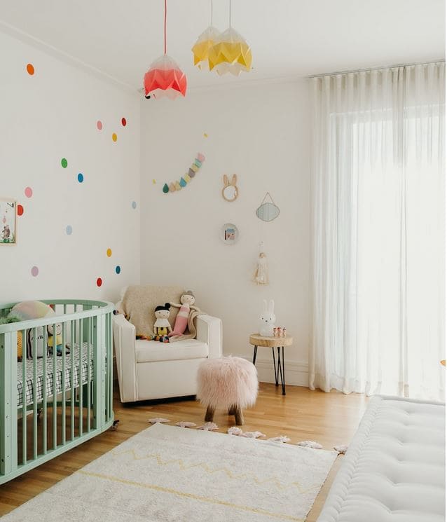 Quarto de bebê em tons claros, berço verde claro e poltrona branca, decorado com circulos pequenos e coloridos nma parede e luminária colorida no teto