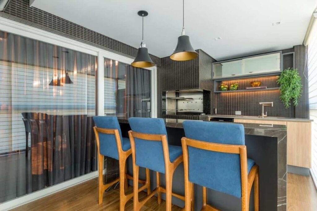 Varanda gourmet com acabamentos em cinza, churrasqueira ao lado do vidro que separa a sala e, em primeiro plano, 3 cadeiras altas com estofado azul