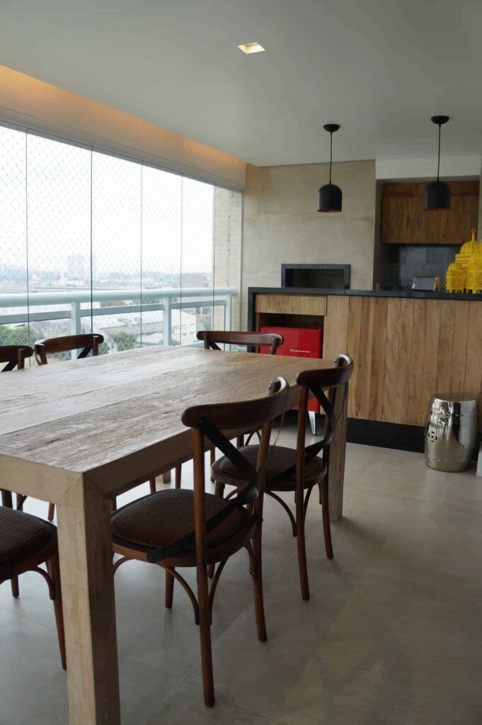 Varanda gourmet com acabamentos em madeira, com mesa e cadeiras também em madeira e à esquerda, vista do apartamento