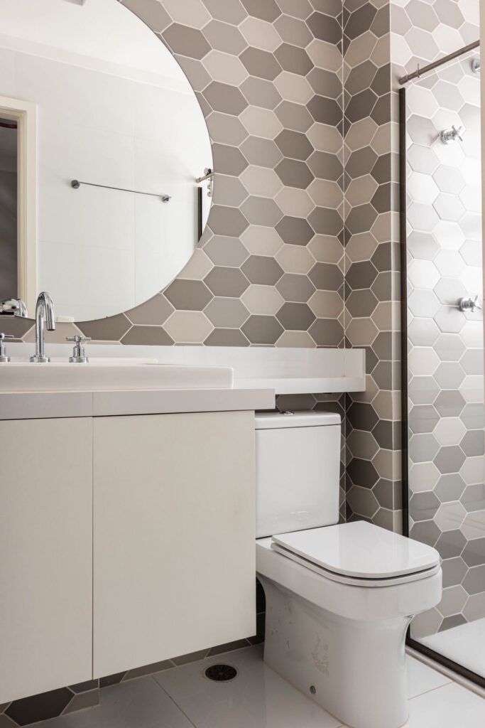 Banheiro pequeno com parede comm azulejo decorativo em formatos de hexágonos em tons de cinza, espelho redondo e louças brancas