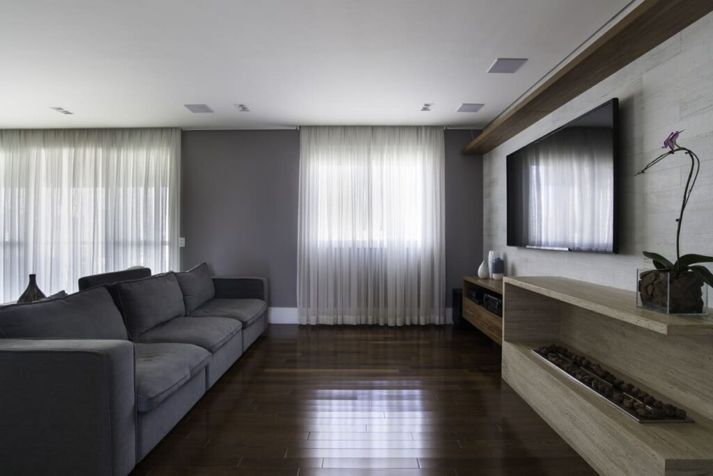 Sala com paredes cinzas e cortinas claras, e piso de madeira escura. Nas laterais, sofá em cinza e estande com TV em madeira de tom mais claro