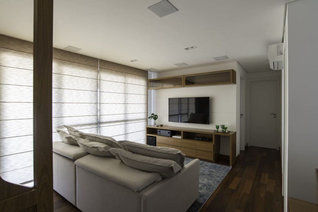 Sala de TV com persiana romana. Sofá e  paredes em tons claros, com TV fixa à parede e móveis e piso em madeira