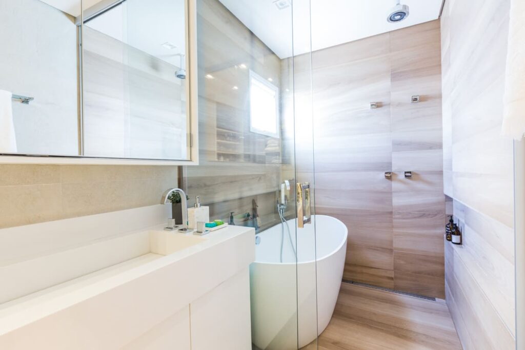 Banheiro com banheira contemporânea integrada ao chuveiro. Banheira e bancada em branco, com acabamentos em porcelanato marrom que imita madeira nas paredes e piso
