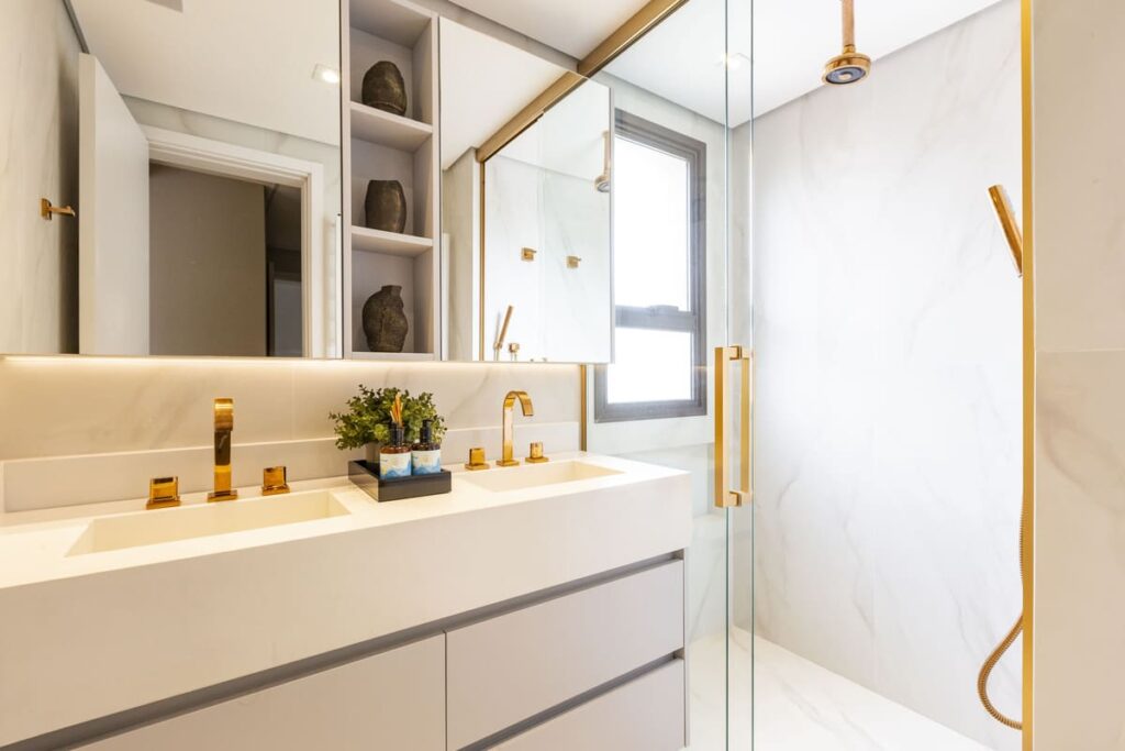 Decoração moderna para banheiro com paredes, louças e gabinete em cores claras e acabamentos como torneiras em metal rose gold. Plantas pequenas na bancada completam a decoração