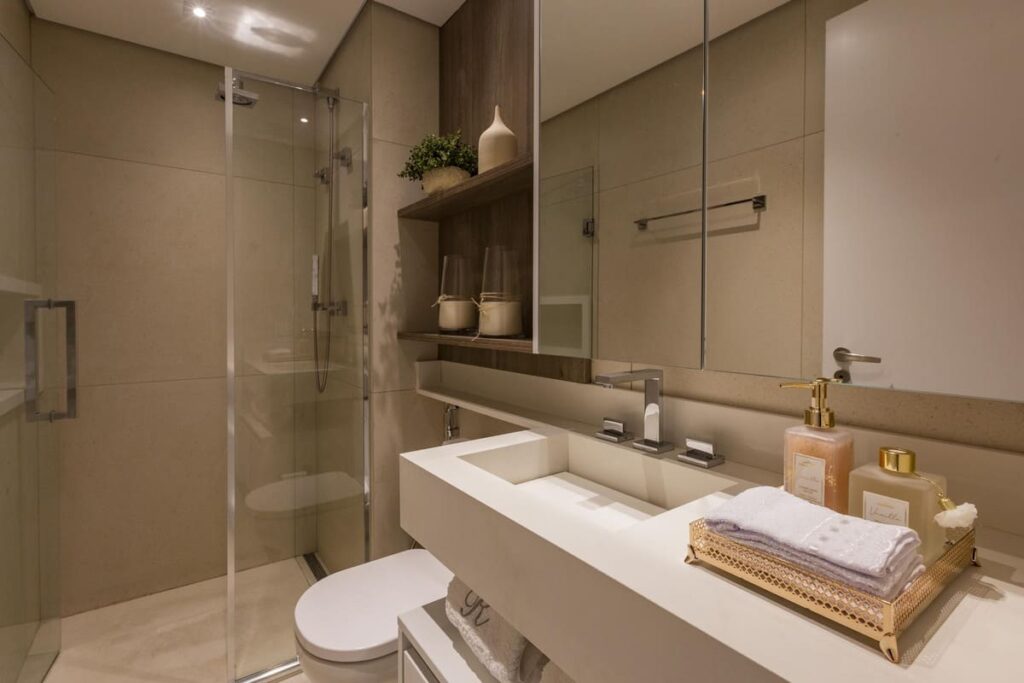 Banheiro com cuba embutida, espelheira e painel decorativo de MDF. Chuveiro com box transparente, espelhos e, na bancada, kit de saboneteira, aromatizador e toalhas de mão