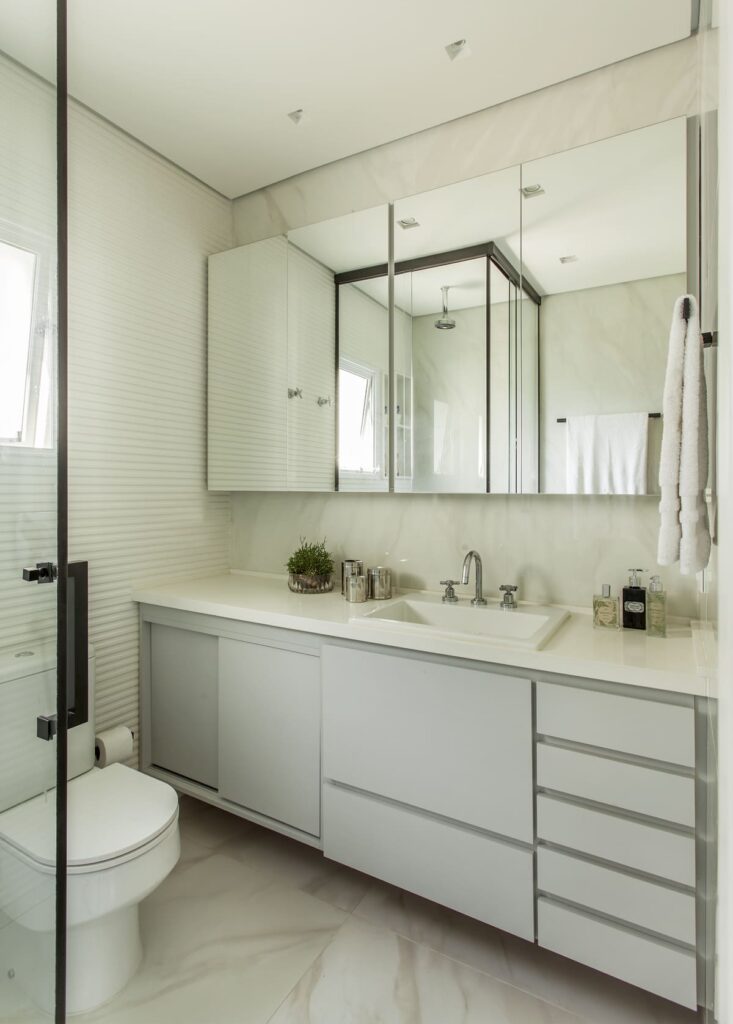 Banheiro em tons de branco, com cabinete em cinza claro. Espelho retangular e um mini vaso de plantas completam a decoração