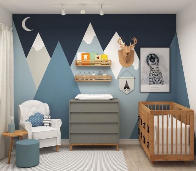 Decoração quarto bebê emnino com tema montanha. Na parede ao fundo, em tons de azul, lua, e montanhas nevadas de várias alturas. Berço em marrom claro, cômoda em cinza e poltrona branca com almofada azul