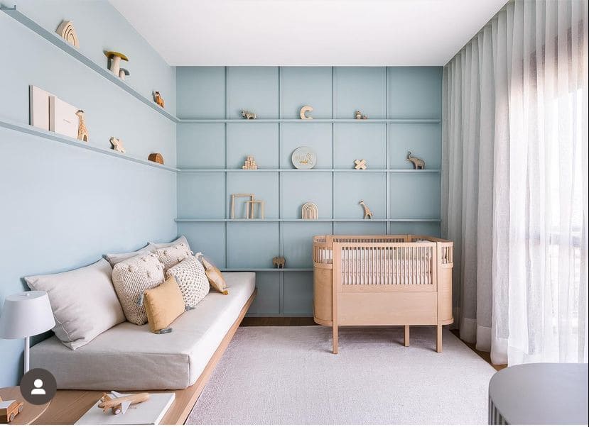 Decoração quarto bebê menino com paredes em azul claro, com prateleiras e objetos de decoração. Cortina de decido clara, com sofá e berço em tons de bege