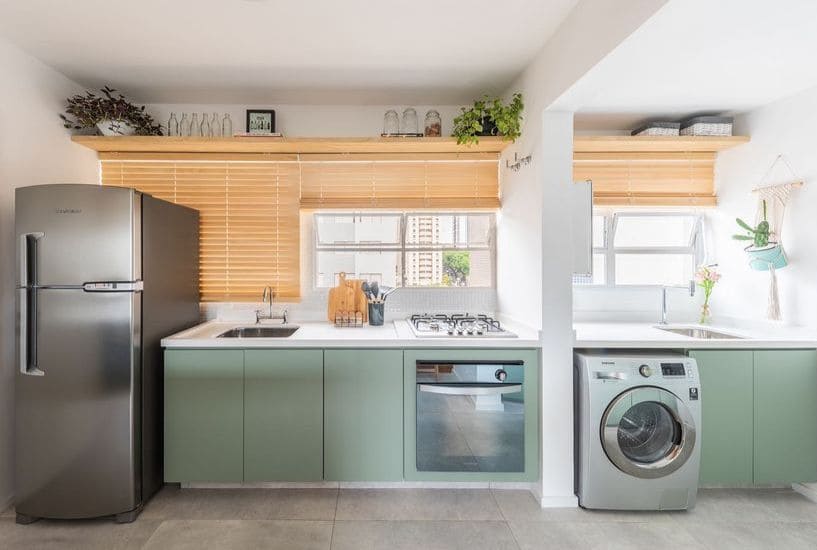 Cozinha e lavanderia separadas por uma parede, mas com mesmos materiais e móveis, em tom de verde claro e bancada branca, com eletrodomésticos em inox. Janela ao fundo com persiana em madeira clara